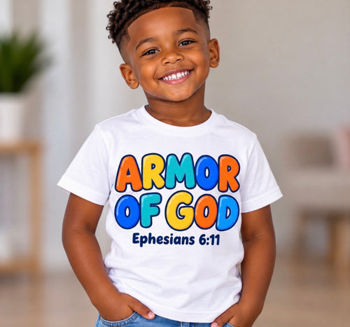 A young boy smiling and wearing a white graphic t-shirt with the text 'ARMOR OF GOD' in colorful letters and 'Ephesians 6:11' underneath, set against a blurred indoor background.